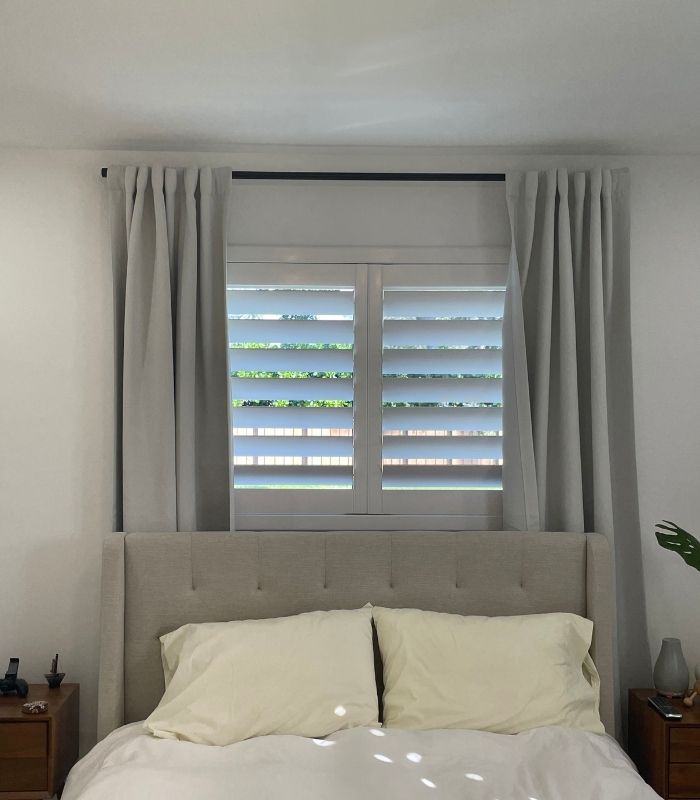 Full height plantation shutters mounted inside a bedroom window frame with decorative curtains on each side, providing privacy and natural light control.